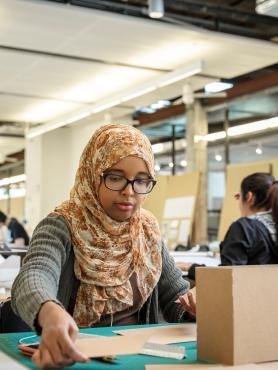 Female student wearing headscarf works with cardboard model at her desk in studio