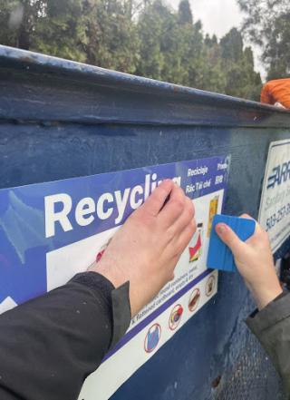 Two hands applying a large blue recycling sticker to a blue dumpster