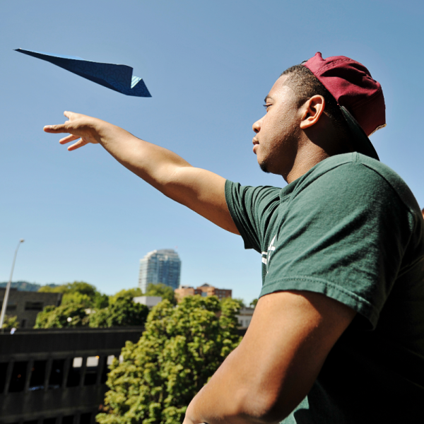 Photo of student wearing a red baseball cap and green t-shirt tossing a paper airplane into the air.