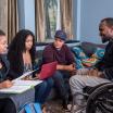 4 people of color sitting down and collaborating together, three on a couch and one in a wheelchair