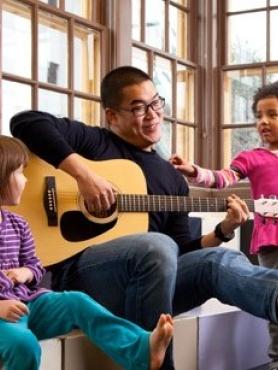 Two happy young children surrounding an early childhood educator playing a guitar. Sunlight pours into room through large windows.