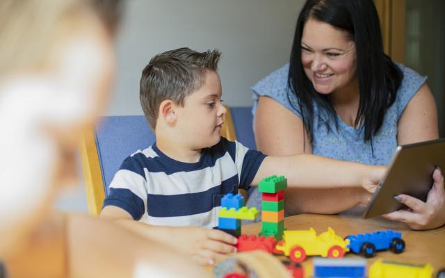 A young boy plays with blocks as a dark-haired woman smiles at him encouragingly.