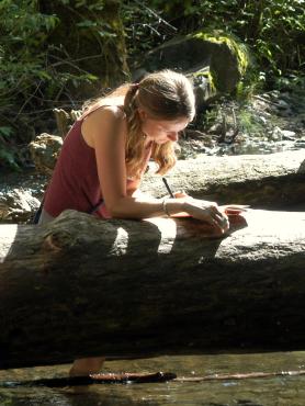 Man and woman sitting on log by stream, writing in notebooks