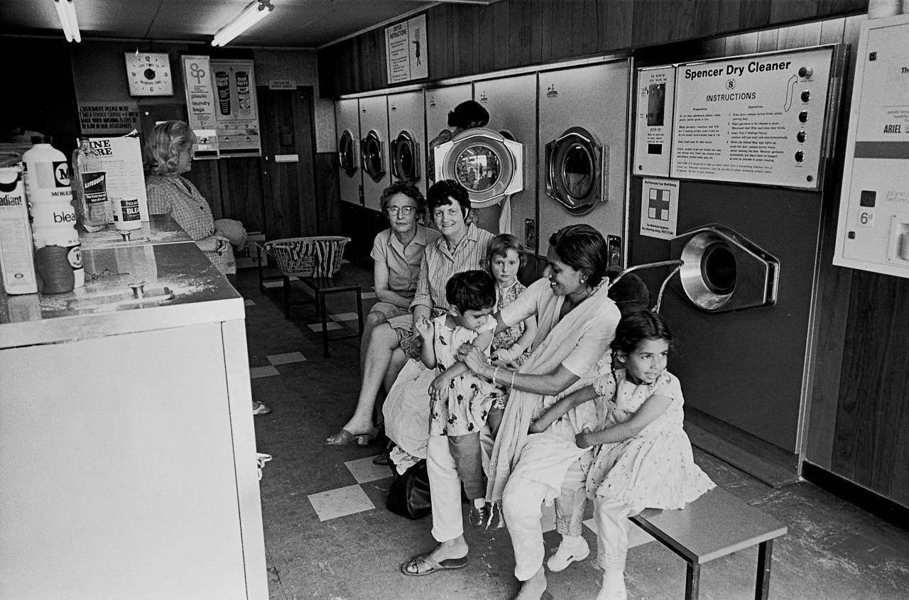 In a Bradford laundrette, Nick Hedges, 1970. British women and Indian mother and children on a laundry bench in Bradford, England