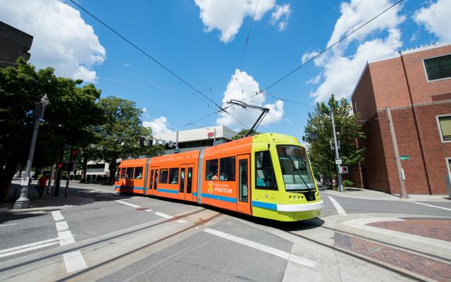 The orange and green streetcar crosses the street, diagonally across the frame. The sky is blue and there are a few clouds in the sky behind. 
