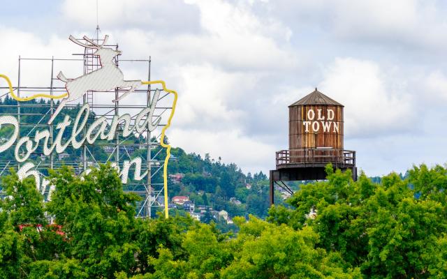 On the left side of the photo is the Portland city sign, and to the right is a brown water tower that says "Old Town". There are trees in the foreground and background.  