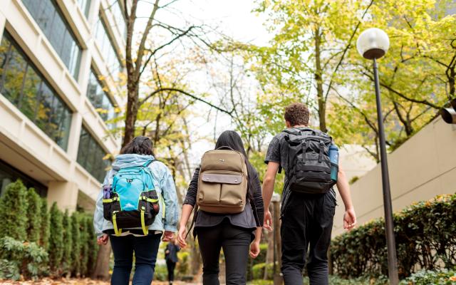 Three students wearing backpacks walk on a path between trees and buildings with their back turned to the camera.