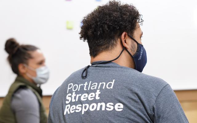 Photo of person facing away from the camera wearing a mask and a grey shirt that says "Portland Street Response".
