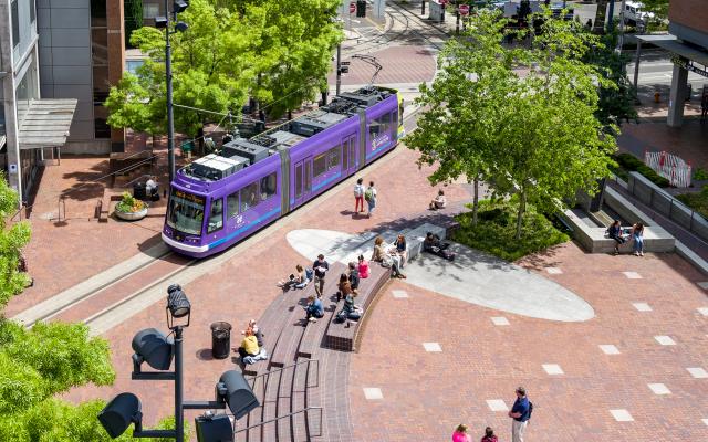 Purple streetcar going through the PSU urban center. There are people around and the trees are green.