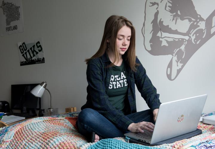 PSU student sitting on a bed in a dorm room and working on a laptop