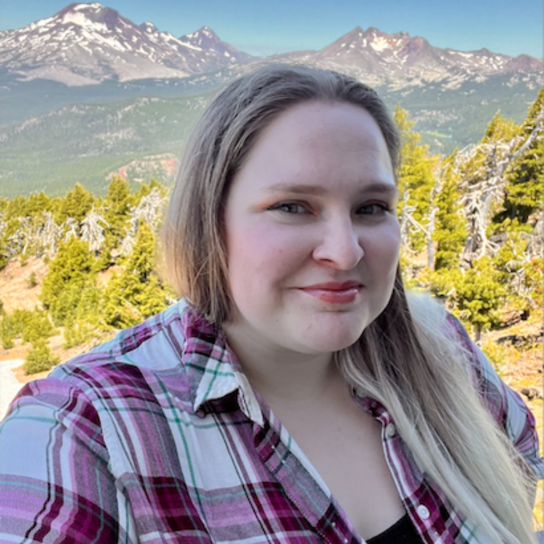 Female-identified person with long, blonde hair wearing a plaid shirt with mountains in the backround.