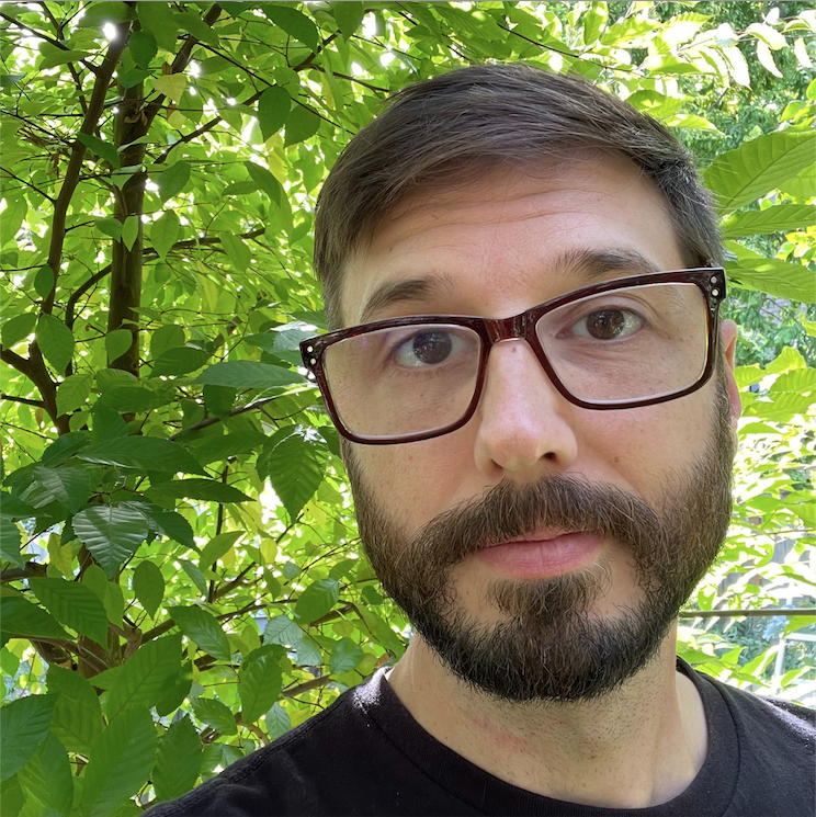 Person with short, brown hair, beard, wearing a black shirt, with trees in the background.