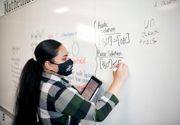 PSU student with light skin and long dark hair pulled back in a ponytail. They're using a whiteboard to work on an equation. They have on a black face mask with white writing and a green, white, and black plaid long sleeve shirt.