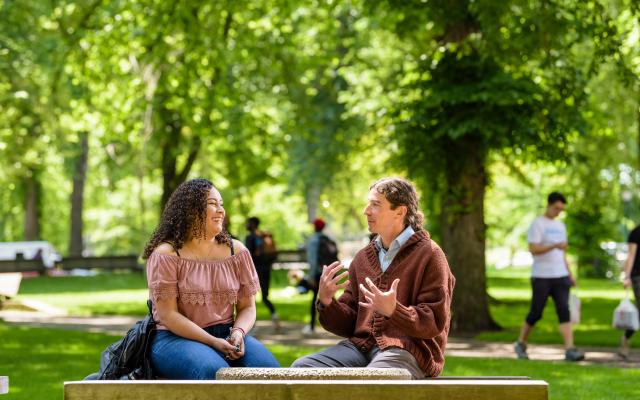 Photo of two students on a bench in the park blocks have a conversation