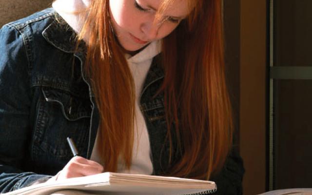 Photo of a student sitting against a wall writing notes in her booklet