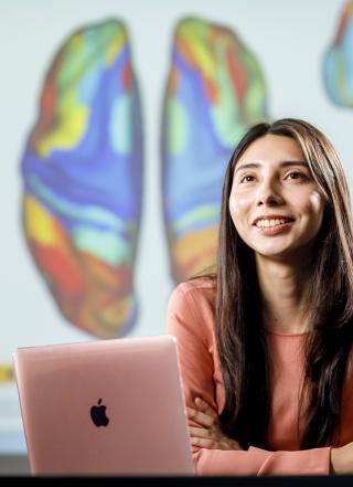 Smiling woman with long dark hair and light images of a brain. The brains are multicolored skin seated with arms folded behind a pink laptop. Behind the woman is a screen with multiple brains. The brains are multicolored, with each color representing a different part of the brain.