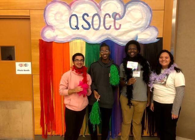 4 QTBIPOC stand in front of a cloudy rainbow QSOCC sign