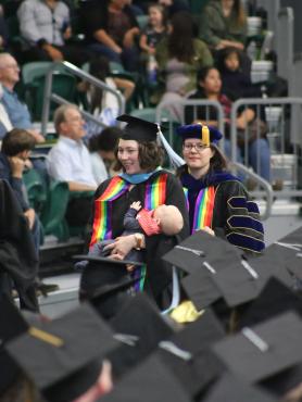 three graduates wear rainbow stolls. one holds a baby