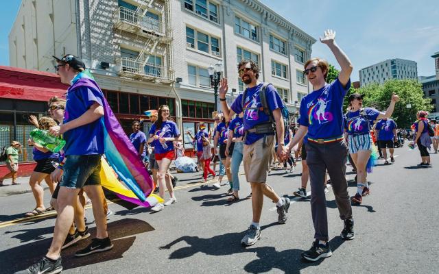a group of PSU affiliates walk in the Portland Pride parade in matching purple shirts. A white queer couple holds hands and waves