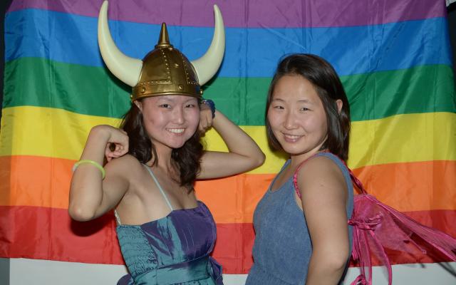 two asian student smile at the camera in front of a rainbow flag. one flexes their bicepts with a viking hat on, the other stands in super hero pose, smiling strongly