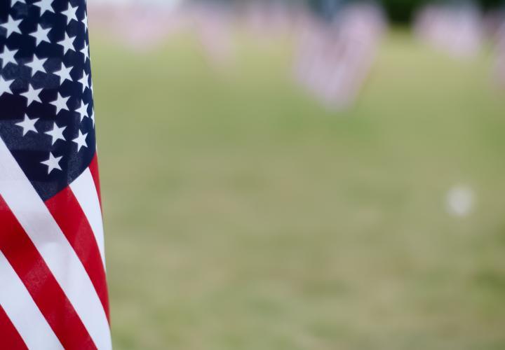 American flag in the foreground with more American flags blurred out in a grassy field in the background.