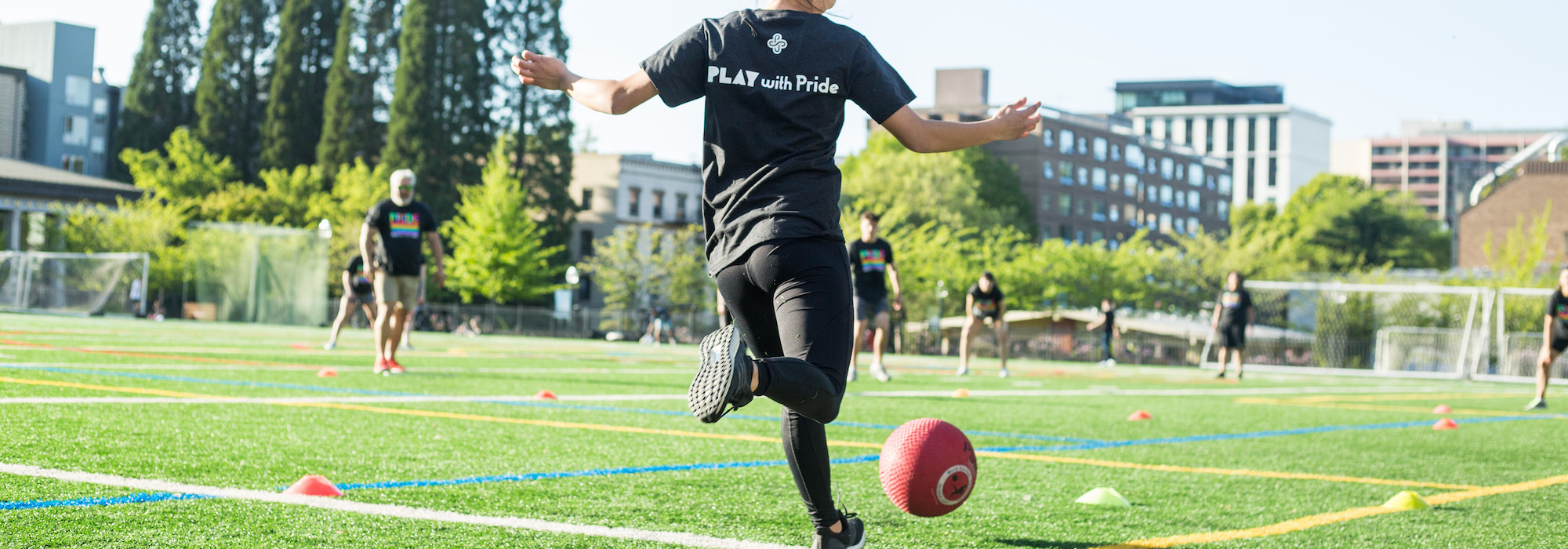 Person with a "Play with Pride" shirt is kicking a ball to teammates during the Pride Kickball Tournament in 2019 at Stott Field.
