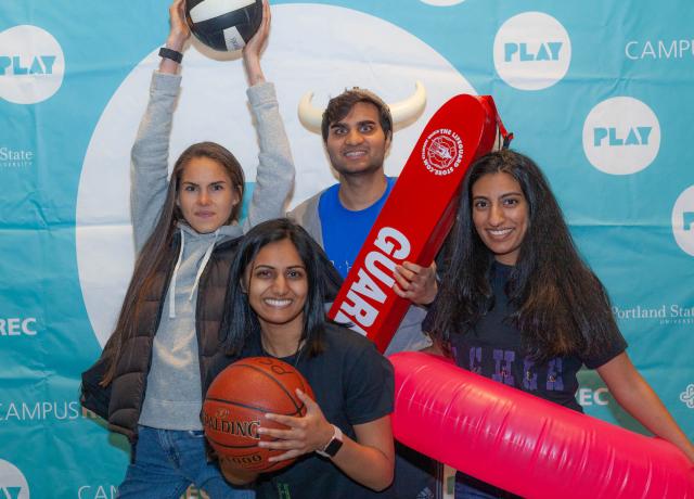 A group of students poses at a photo booth with a volleyball, basketball, inner tube, and lifeguard float at Night at the Rec 2019