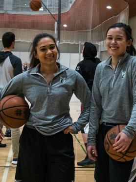Two female basketball players smile while holding the basketball on the court.