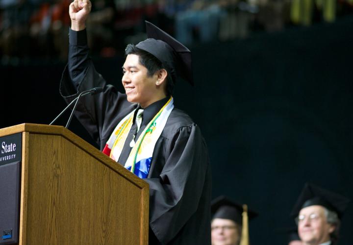 The image is of a person wearing graduation regalia, they are standing at a podium with a fist in the air. 