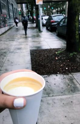 There is a hand holding a cup of coffee above the sidewalk, there are people walking in the background.