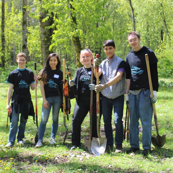Five students stand in a park holding shovels at a service project