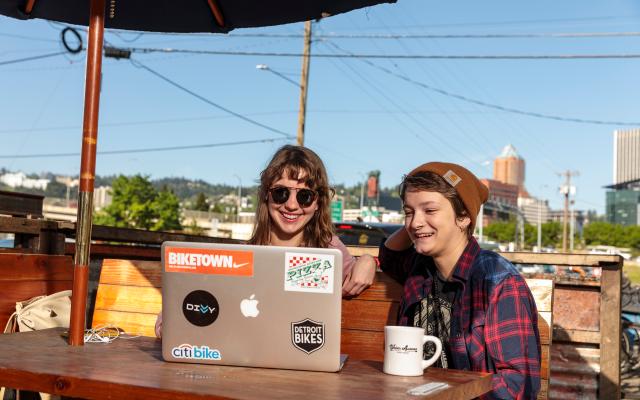two people sitting outside, looking at a laptop, and smiling