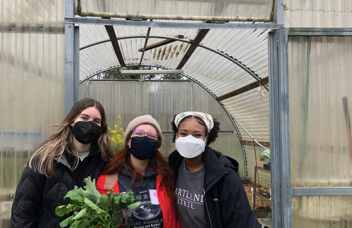 Three students with masks on stand in front of a greenhouse. One student holds a bundle of greens.