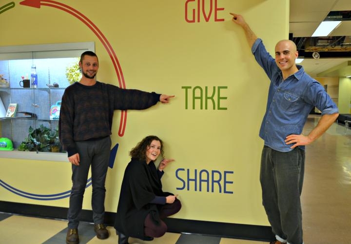 Three people pointing to wall mounted sign saying give take share