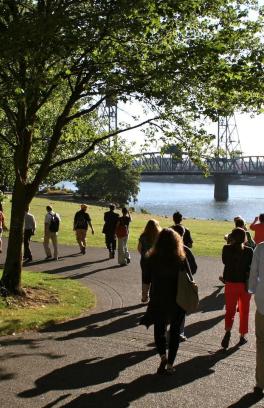 A group of people walk along a path alongside the Willamette River, with the Hawthorne Bridge in the distance