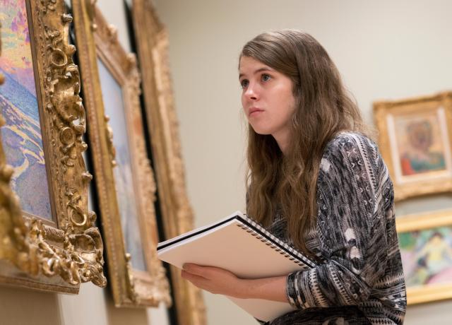 a student holds a notebook and looks at a wall of paintings in ornate gilded frames