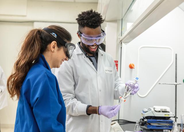two students wearing lab coats and safety goggles are working in a lab