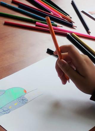 Close-up of a tabletop with a child's hand holding a colored pencil. On a piece of paper is a turquoise drawing of a car on a street. More colored pencils are in the background.