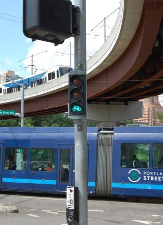 Multimodal transportation: A streetcar passes under a bridge on which a Max train travels, with a bike signal in the foreground.