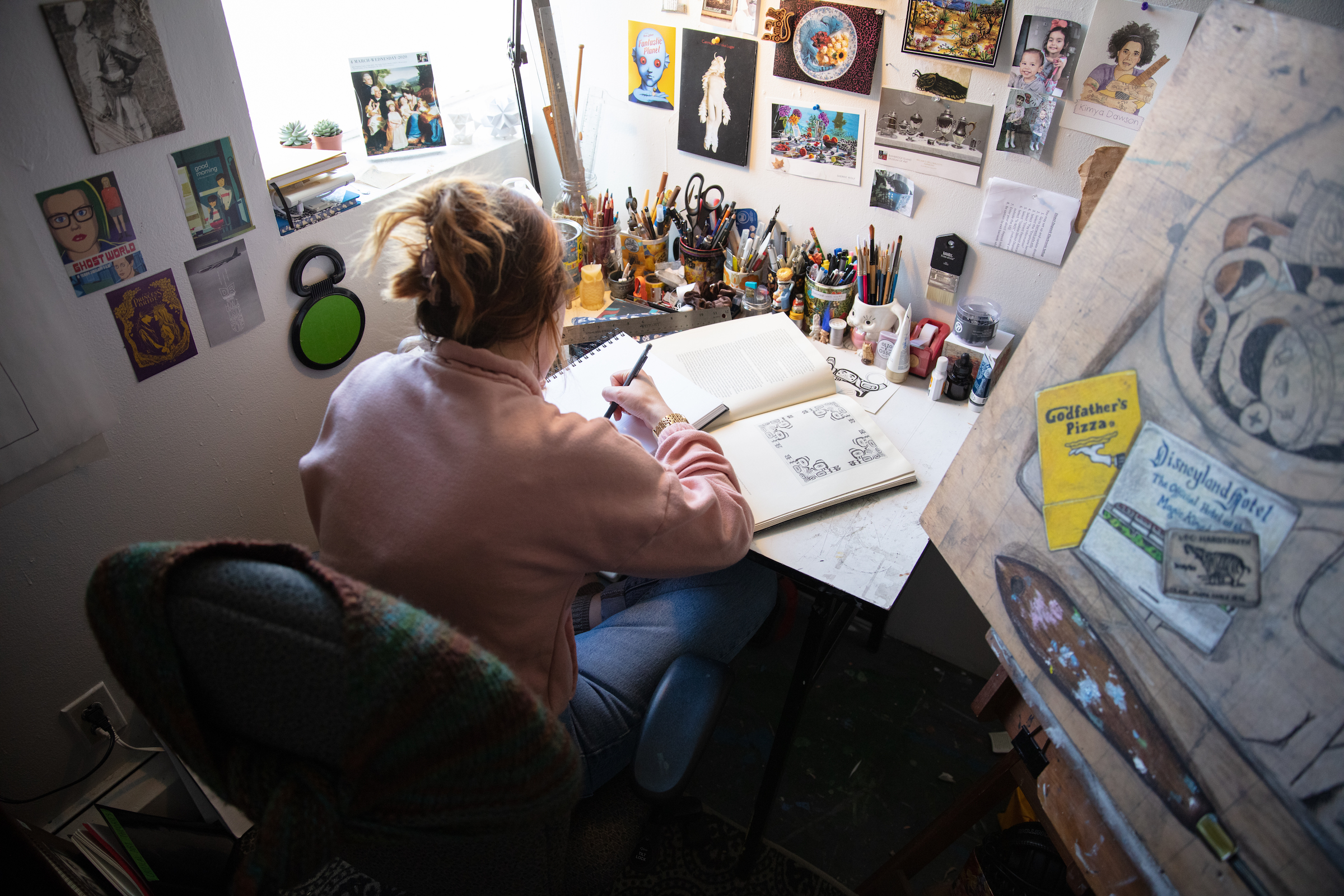 Graduate student working at a drawing table by a window, surrounded by reference images and art supplies