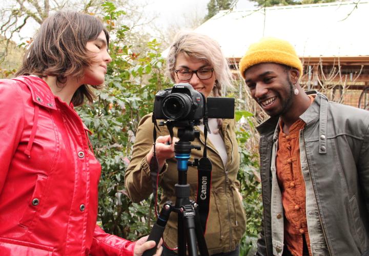 Three students in a video and community course gather around a camera on a tripod