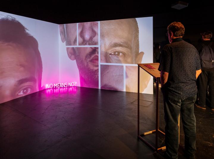 Man stands at a podium in front of a video projection with collage of male faces