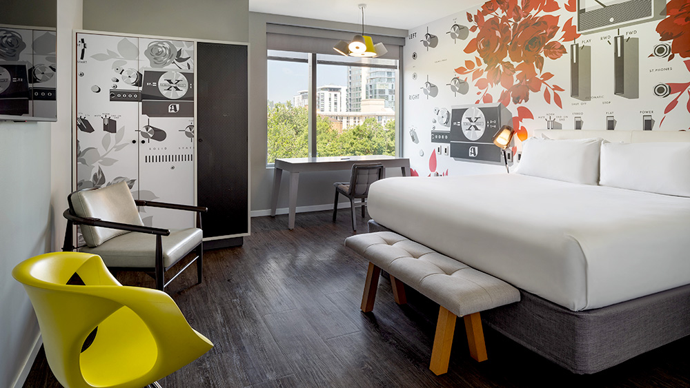 Interior of a hotel room with chairs at left, bed at right, view of the city through a window and mural on the wall.