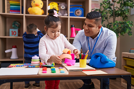 A male teacher sporting an undercut and a light blue button-up crouches down to assist a young girl with plastic learning toys.