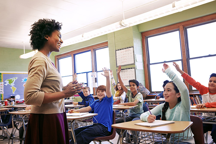 A young Black woman smiles as she addresses a classroom full of children with raised hands.