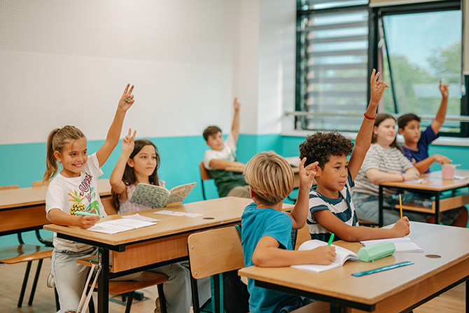 A white and teal classroom is full of students with their hands raised.