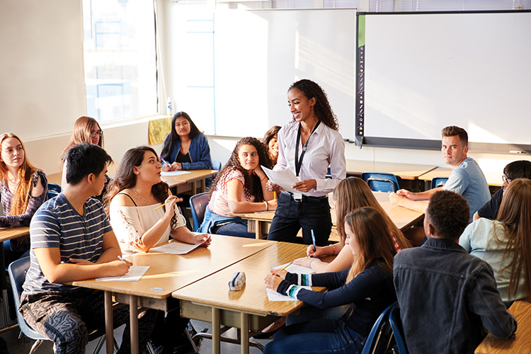 A teacher stands in the middle of a secondary school classroom talking with her students.