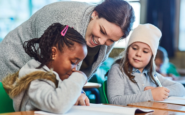 A Caucasian teacher with dark brown hair pulled back assists two students with their classwork.