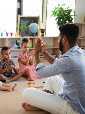 A male teacher with dark, curlly hair and beard sits cross-legged with a couple of children teaching them hand games.