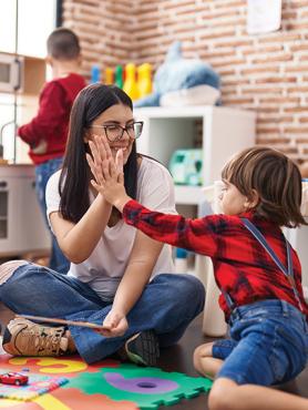 A young woman with long, dark brown hair and glasses high fives a child in a red flannel shirt and denim overalls.